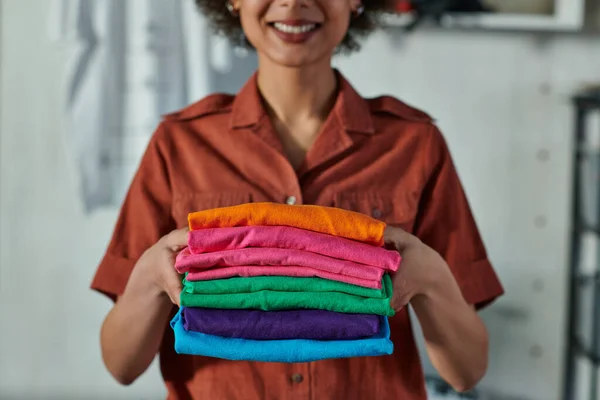 Vista recortada de la borrosa joven y sonriente artesana afroamericana sosteniendo la ropa y de pie en el estudio de impresión, concepto emprendedor centrado en el cliente - foto de stock