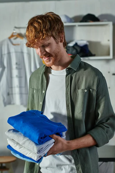 Cheerful young redhead craftsman holding clothes while standing and working in blurred print studio at background, customer-focused entrepreneur concept — Stock Photo