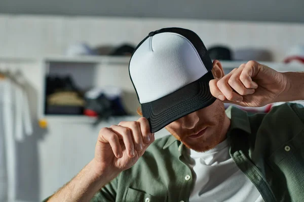 Young craftsman covering face and wearing snapback while standing and working in blurred print studio at background, customer-focused entrepreneur concept — Stock Photo