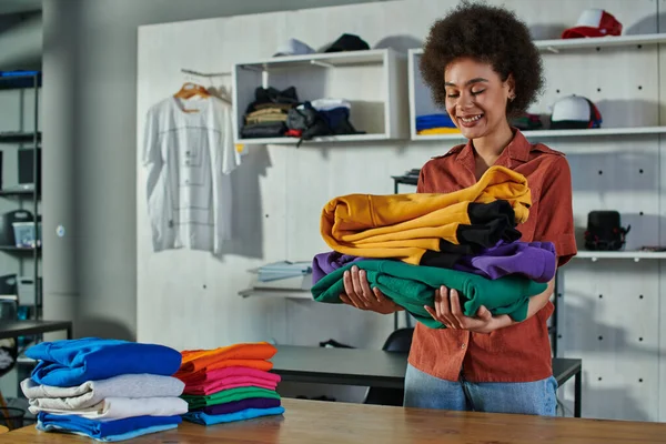 Positive young african amercan craftswoman holding clothes near working table while standing in blurred print studio at background, customer-focused entrepreneur concept — Stock Photo