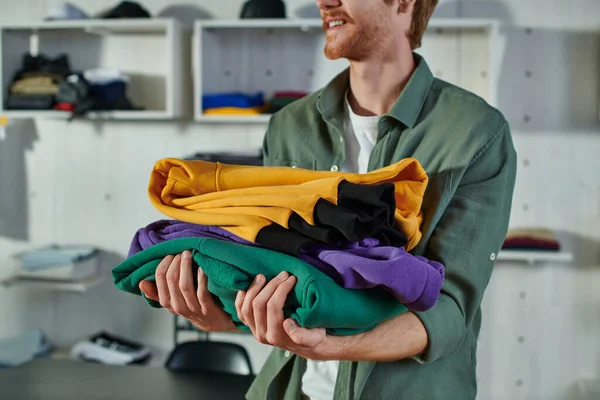 Ausgeschnittene Ansicht eines lächelnden jungen rothaarigen Handwerkers, der Kleidung hält, während er arbeitet und im verschwommenen Druckstudio im Hintergrund steht, kundenorientiertes Unternehmerkonzept — Stockfoto