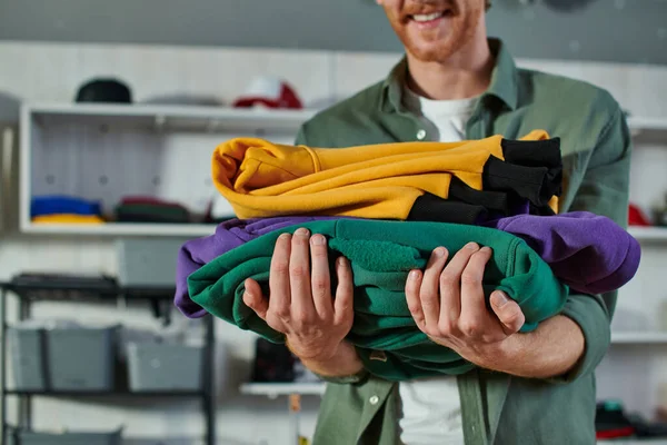Cropped view of blurred cheerful craftsman holding clothes and standing while working in print studio at background, customer-focused entrepreneur concept — Stock Photo
