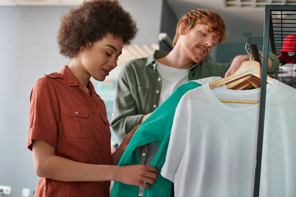 Smiling young african american craftswoman looking at clothes on hangers near blurred redhead colleague and standing together in print studio, young small business owners concept — Stock Photo