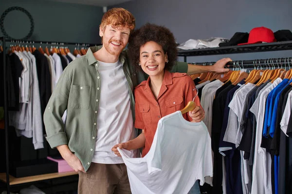Positive young multiethnic craftspeople looking at camera while standing near clothes on hangers and working in print studio, young small business owners concept — Stock Photo