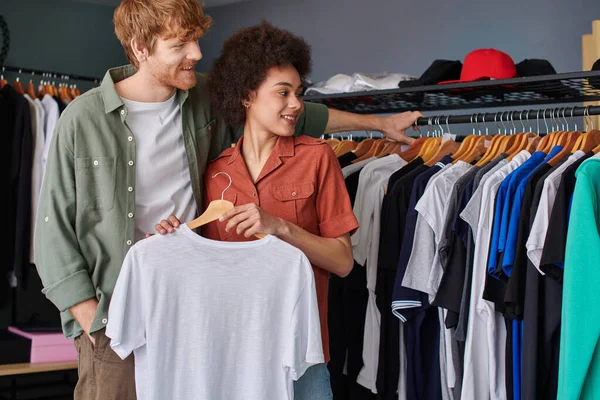 Fröhlicher junger rothaariger Handwerker, der Kleidung auf Kleiderbügeln betrachtet, während er neben einem afrikanisch-amerikanischen Kollegen im Druckstudio steht, junge Kleinunternehmer — Stockfoto