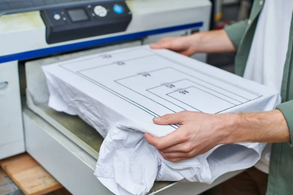 Обрезанный вид молодого ремесленника, держащего футболку с маркировкой возле экранной печатной машины и работающего в типографии, ориентированной на клиента концепции малого бизнеса — стоковое фото