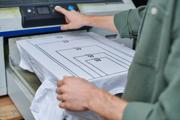 Ausgeschnittene Ansicht eines verschwommenen jungen Handwerkers mit Siebdruckmaschine in der Nähe eines T-Shirts mit Markierung im Druckstudio, kundenorientiertes Kleinunternehmenskonzept — Stockfoto
