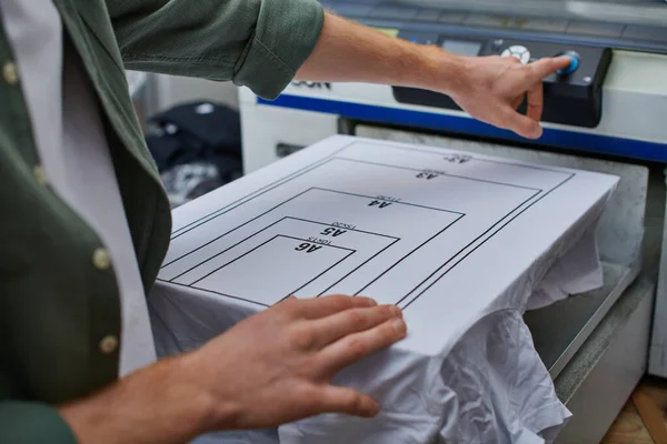 Ausgeschnittene Ansicht junger Handwerker mit verschwommener Siebdruckmaschine in der Nähe von T-Shirts mit Markierung während der Arbeit im Druckereistudio, kundenorientiertes Kleinunternehmenskonzept — Stockfoto