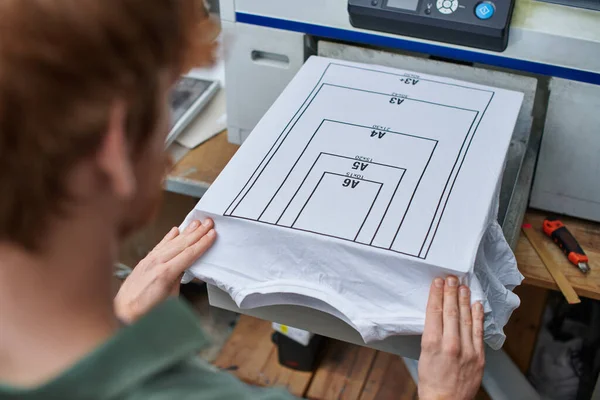 Hohe Blickwinkel auf junge verschwommene Handwerker, die mit T-Shirt mit Markierung und Siebdruckmaschine im Hintergrund im Druckstudio arbeiten, kundenorientiertes Kleinunternehmenskonzept — Stockfoto