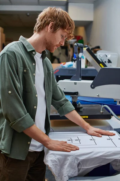 Seitenansicht eines jungen rothaarigen Handwerkers, der mit T-Shirt und Siebdruckmaschine im verschwommenen Druckstudio im Hintergrund arbeitet, kundenorientiertes Kleinunternehmenskonzept — Stockfoto