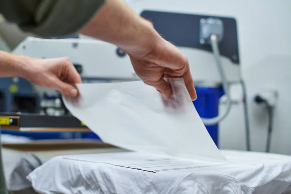 Ausgeschnittene Ansicht junger Handwerker, die Schicht auf T-Shirt legen und im Hintergrund im Druckereistudio auf Siebdruckmaschine arbeiten, kundenorientiertes Kleinunternehmenskonzept — Stockfoto