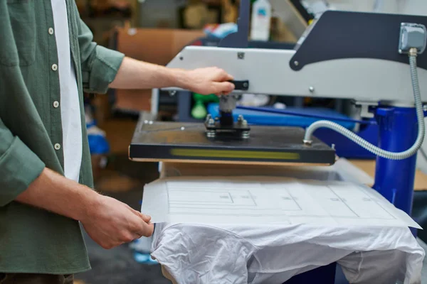 Ausgeschnittene Ansicht des Handwerkers, der Schicht auf T-Shirt hält und mit Siebdruckmaschine in verschwommener Werkstatt arbeitet, kundenorientiertes Kleinunternehmenskonzept — Stockfoto