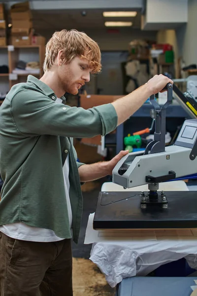 Вид сбоку молодой рыжеволосый ремесленник, работающий с трафаретной печатной машиной и футболкой в размытой типографии на заднем плане, ориентированная на клиента концепция малого бизнеса — стоковое фото
