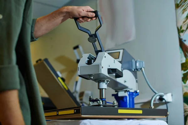 Ausgeschnittene Ansicht eines jungen Handwerkers mit Siebdruckmaschine auf Kleidung, während er im verschwommenen Druckstudio im Hintergrund arbeitet, kundenorientiertes Kleinunternehmenskonzept — Stockfoto