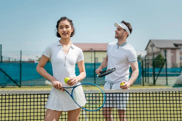 Счастливая женщина в активной одежде проведение теннисной ракетки рядом с мужчиной, теннисисты на корте — стоковое фото