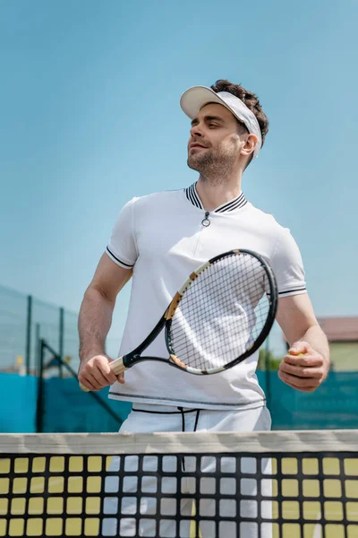 Hombre feliz en visera deportiva y desgaste activo celebración de raqueta de tenis y de pie cerca de la red en la cancha - foto de stock