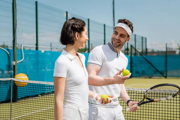 Positive man and woman holding tennis balls and racket on court, hobby and leisure — Stock Photo