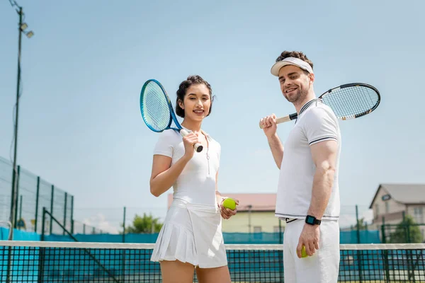 Счастливые мужчина и женщина в спортивной одежде стоя с теннисными ракетками и мячами на корте, глядя в камеру — стоковое фото