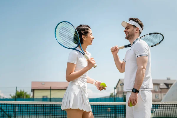 Счастливые мужчина и женщина в спортивном чате, стоя с теннисными ракетками и мячами на корте — стоковое фото