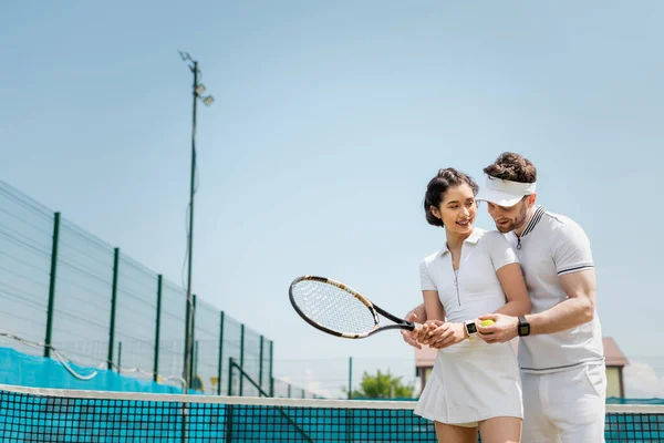 Счастливый мужчина учит подругу играть в теннис на корте, держа рейнджеры и мяч, спорт и веселье — стоковое фото
