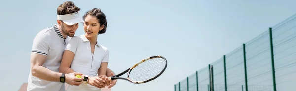 Знаменосный, жизнерадостный мужчина учит подругу играть в теннис на корте, держа в руках рейнджеры и мяч — стоковое фото