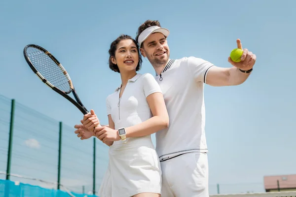 Happy man pointing away near girlfriend on tennis court, holding rackets , sport and romance — Stock Photo