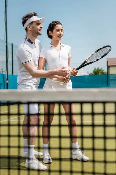 Счастливый мужчина учит подругу играть в теннис на корте, держа рейнджеры, теннисную сетку, летние виды спорта — стоковое фото