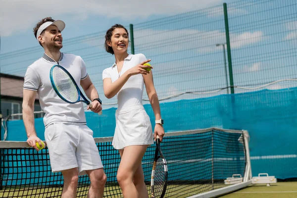 Mujer feliz señalando lejos, hombre sonriendo y sosteniendo raqueta de tenis, verano, deporte de pareja, pasatiempo - foto de stock
