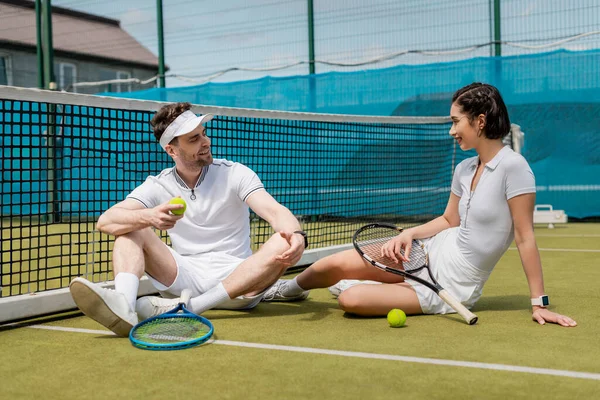 Feliz hombre y mujer en ropa deportiva sentado en la cancha de tenis, raquetas y pelotas, verano, deporte de pareja - foto de stock