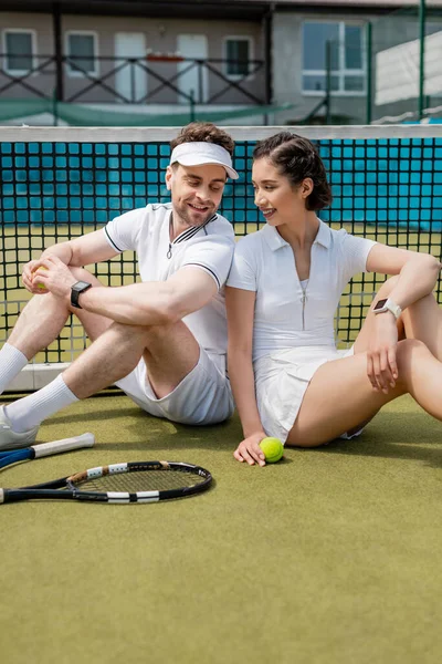 Pareja positiva sentada cerca de la red de tenis, raquetas y pelota, actividad de verano, ocio y diversión - foto de stock