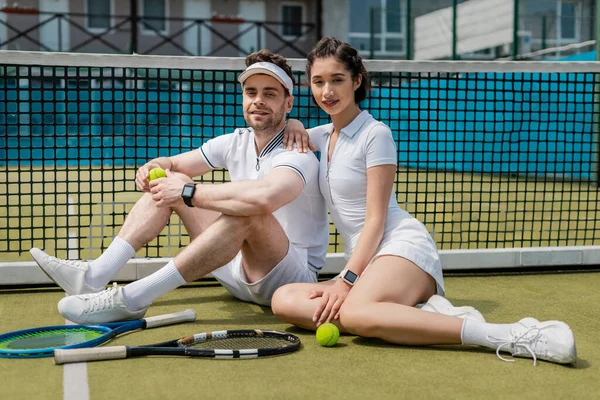 Позитивный мужчина и женщина, сидящие рядом теннисные сетки, ракетки и мяч, летняя деятельность, отдых и развлечения — стоковое фото