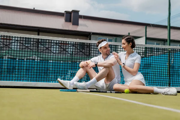 Estilo de vida saludable, hombre y mujer alegres sentados cerca de la red de tenis, raqueta y pelota, positividad - foto de stock