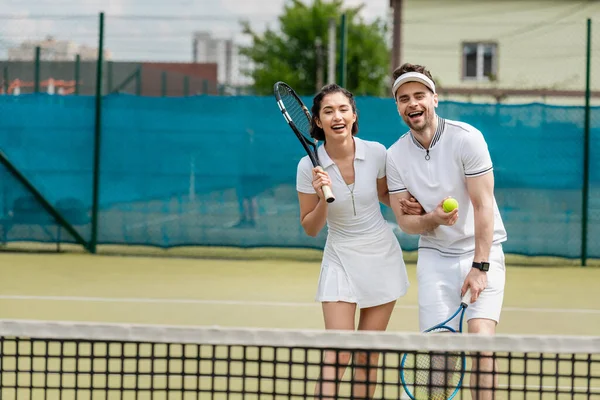 Cheerful sporty couple in active wear holding rackets and ball near tennis net, hobby and sport — Stock Photo