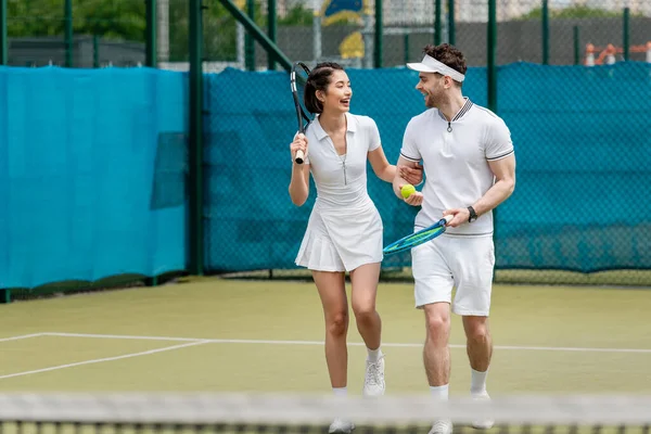 Счастливая спортивная пара в активной одежде ходьба по теннисному корту, ракетки и мяч, хобби и спорт — стоковое фото