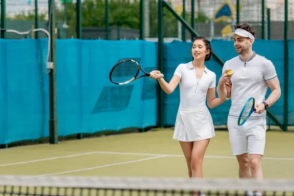 Счастливая женщина с теннисной ракеткой рядом с бойфрендом на корте, романтика и спорт, лето — стоковое фото