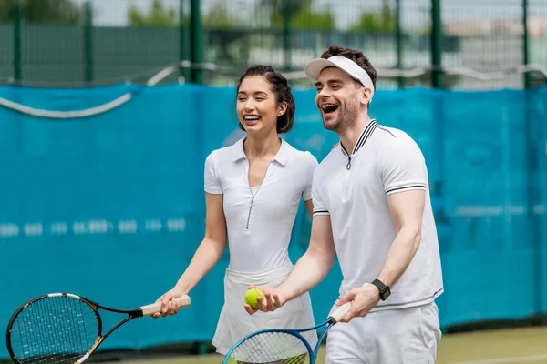 Веселая пара в активной одежде смеется на теннисном корте, игроки, ракетки и мяч, спорт — стоковое фото