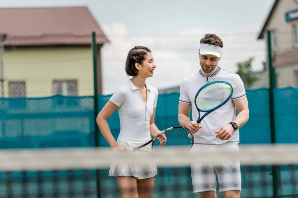 Весёлый мужчина в активной одежде смотрит на теннисную ракетку, счастливая женщина улыбается на теннисном корте, спорт — стоковое фото