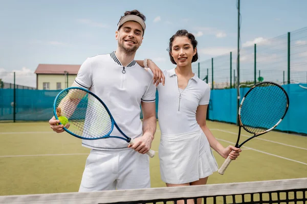 Счастливая спортивная пара в активной одежде глядя на камеру на теннисном корте, хобби и спорт, лето — стоковое фото