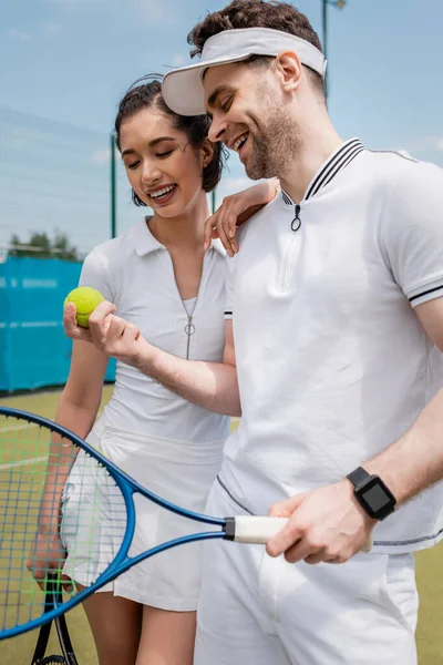 Casal alegre em desgaste ativo olhando para bola de tênis na quadra, lazer e esporte, diversão de verão — Fotografia de Stock