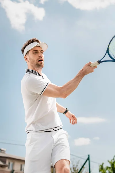 Hombre guapo en gorra de visera y ropa activa que sostiene la raqueta en la pista de tenis, revés, pasatiempo y deporte - foto de stock