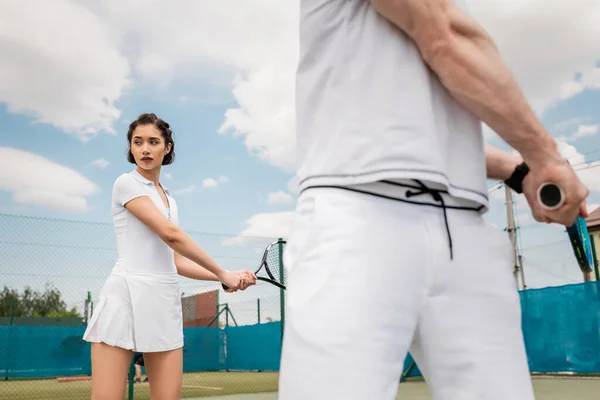 Привлекательная женщина держит теннисную ракетку возле бойфренда на размытом переднем плане, теннисный корт, спорт — стоковое фото