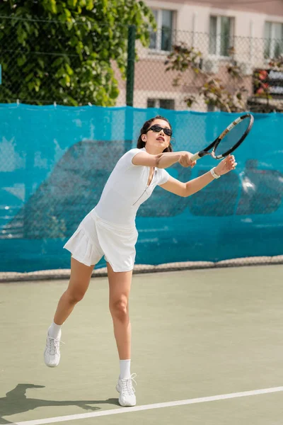 Целенаправленная женщина в солнцезащитных очках и активной ношении ракетки во время игры в теннис на корте, спорт — стоковое фото