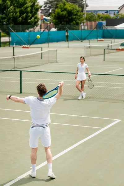 Hombre golpeando pelota de tenis con raqueta, mujer atlética de pie en la cancha de tenis, los jugadores en uso activo - foto de stock