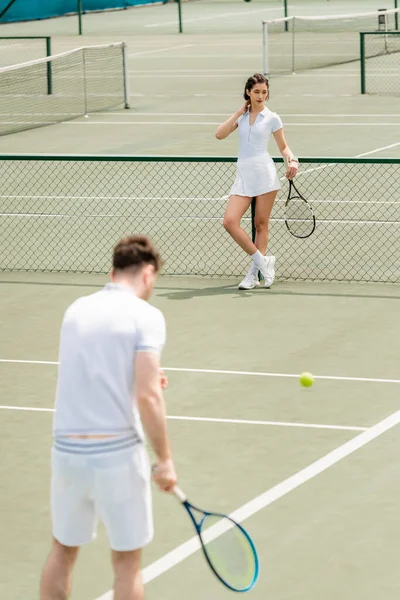 Woman standing near tennis net and holding racket, man in active wear on blurred foreground — Stock Photo