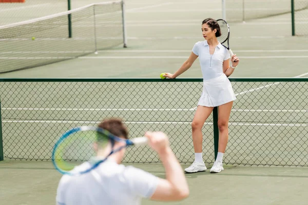 Joueuse de tennis debout près du filet et tenant la raquette, homme en tenue active au premier plan flou — Photo de stock