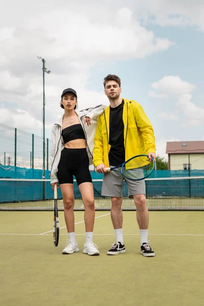 Мужчина и женщина в стильной активной одежде стоят на корте с теннисными ракетками, здоровый образ жизни, спорт — стоковое фото