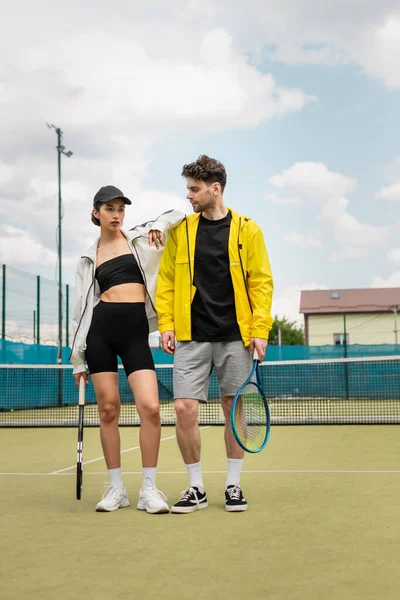Coppia alla moda in piedi sul campo da tennis con racchette, abbigliamento sportivo alla moda, tennisti — Foto stock