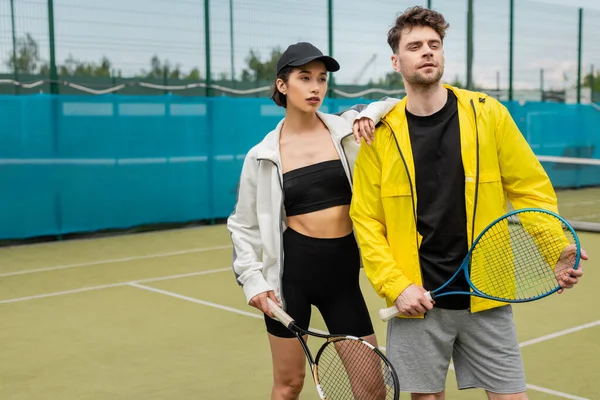 Deporte, pareja de moda de pie en la cancha con raquetas de tenis, hombre y mujer en trajes elegantes - foto de stock