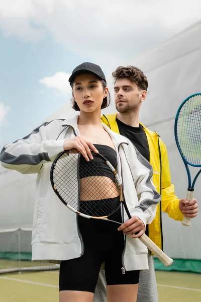 Модные мужчина и женщина, стоящие на корте с теннисными ракетками, стильная пара, спорт и хобби — стоковое фото
