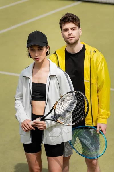 Спорт и стиль, модный мужчина и женщина в спортивной одежде проведение теннисных ракетки на корте — стоковое фото